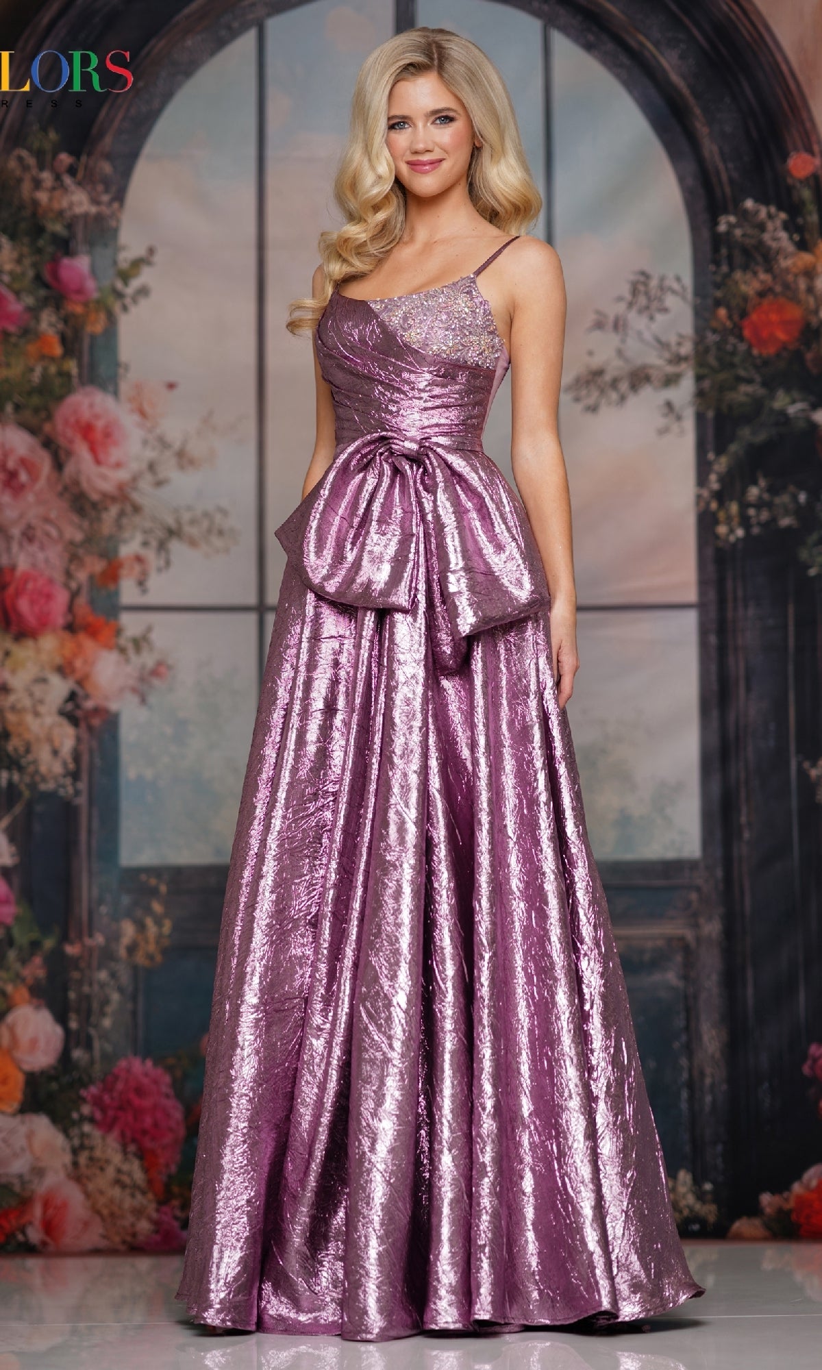 A woman wears the Colors Dress 3793, a sleeveless metallic lavender A-line long prom dress with a large waist bow, standing before a floral and arched window backdrop.
